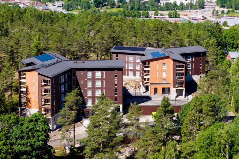 Ilmakuva Kirkkonummen Juhlakallion vähähiilisestä puukerrostalokohteesta: kolme keskikokoista punertavan ja tummanruskeaa puupintaista asuinkerrostaloa metsän reunassa, parvekkeita, aurinkopaneeleja katoilla sekä piha-alue ja ajotie etualalla.