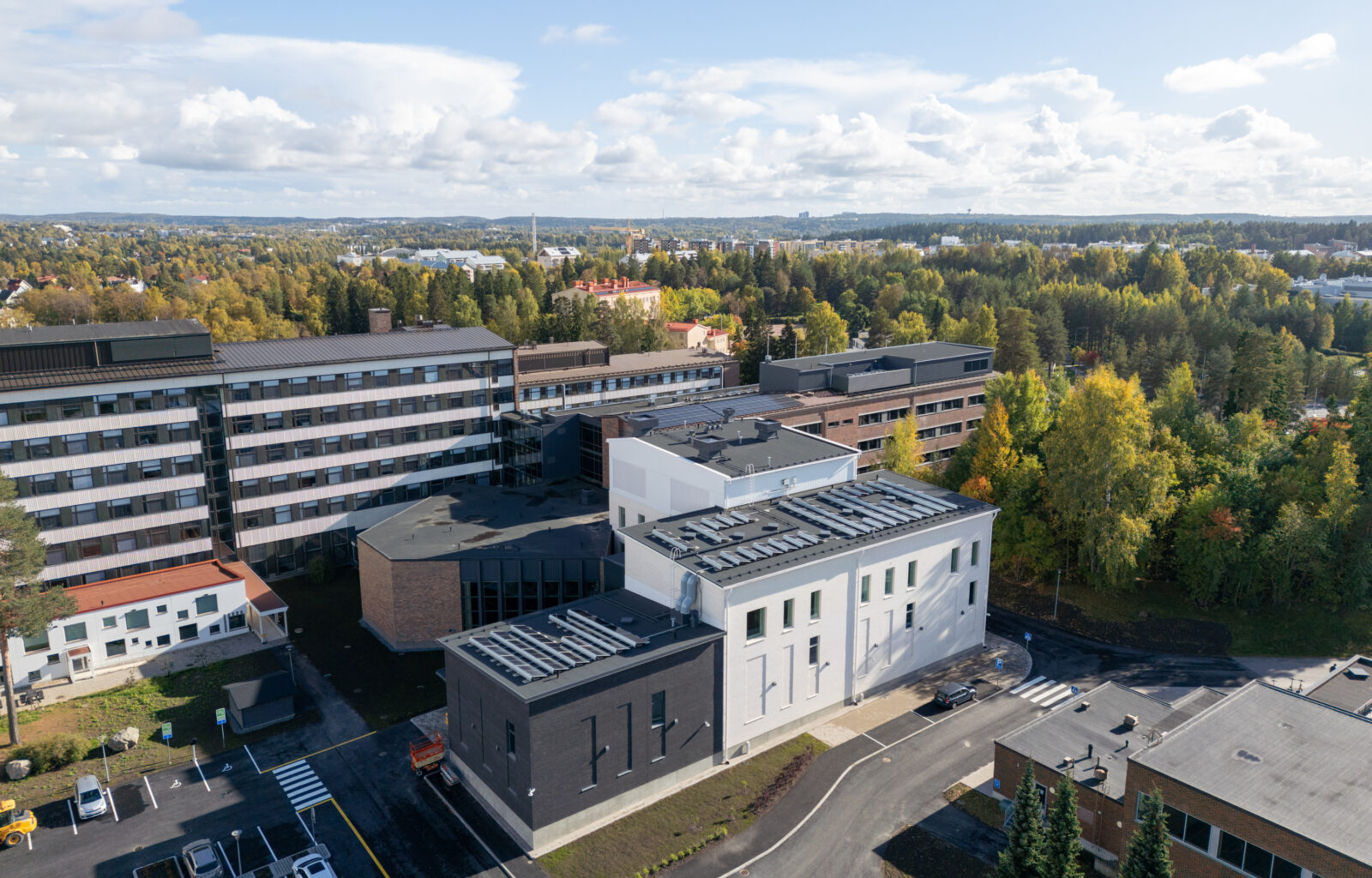 Ilmakuva Tampereen ammattikorkeakoulun Musiikkitalon laajennuksesta ja peruskorjatusta osasta: etualalla moderni valko‑tummanharmaa rakennus, taustalla vanhempi kampusrakennus, pysäköintialue ja kadut sekä syksyiset puut ja kaupunkimaisema horisontissa.