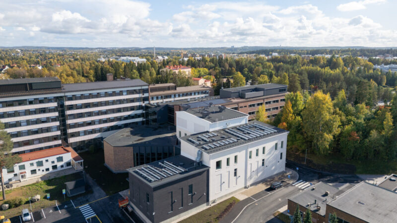 Ilmakuva Tampereen ammattikorkeakoulun Musiikkitalon laajennuksesta ja peruskorjatusta osasta: etualalla moderni valko‑tummanharmaa rakennus, taustalla vanhempi kampusrakennus, pysäköintialue ja kadut sekä syksyiset puut ja kaupunkimaisema horisontissa.