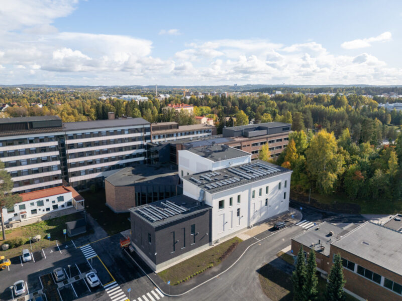 Ilmakuva Tampereen ammattikorkeakoulun Musiikkitalon laajennuksesta ja peruskorjatusta osasta: etualalla moderni valko‑tummanharmaa rakennus, taustalla vanhempi kampusrakennus, pysäköintialue ja kadut sekä syksyiset puut ja kaupunkimaisema horisontissa.