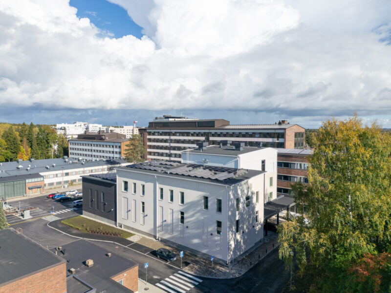Ilmakuva Tampereen ammattikorkeakoulun kampuksesta: etualalla vaalea, moderni suorakaiteen muotoinen Musiikkitalon laajennus, taustalla laajempia kampusrakennuksia ja pysäköintialue, ympärillä syksyisiä puita ja pilvinen taivas.