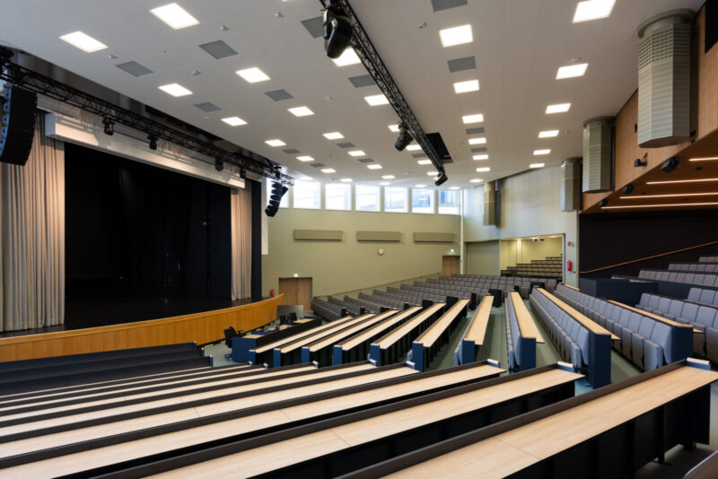 Sisäkuva TAMK Musiikkitalon suuresta modernista auditorosta, jossa vasemmalla lava esirippuineen, katossa valot ja äänentoistolaitteet sekä useat porrastetut penkkirivit pitkine pulpettipöytineen.